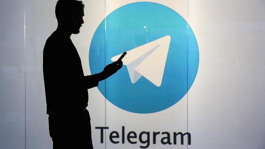 A man is seen as a silhouette as he checks a mobile device whilst standing against an illuminated wall bearing Telegram's logo.