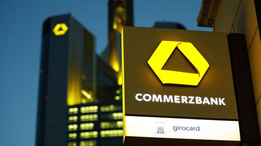 The Commerzbank AG logo sits on an illuminated sign outside a bank branch as the bank's headquarters stand beyond at dusk in Frankfurt, Germany, on Monday, Feb. 5, 2017. 